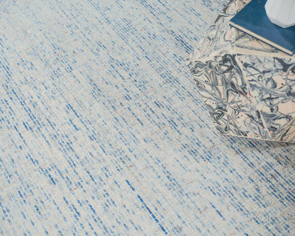 A light blue and ivory hand-tufted rug with subtle blue streaks, beside a marble-patterned cube ottoman and a blue notebook.