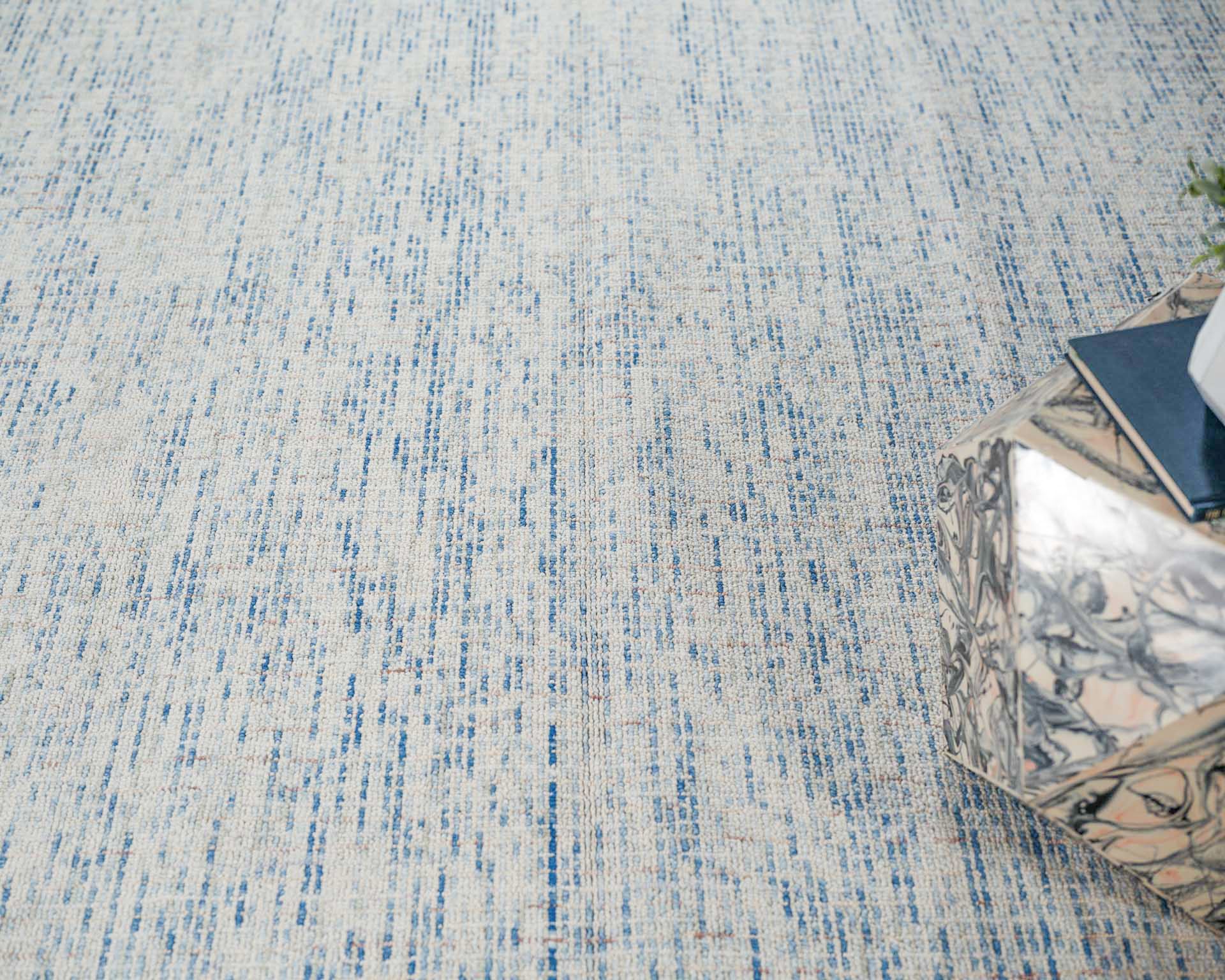 Blue and ivory textured rug with abstract vertical flecks; marble-patterned cube side table with a blue book and small plant.