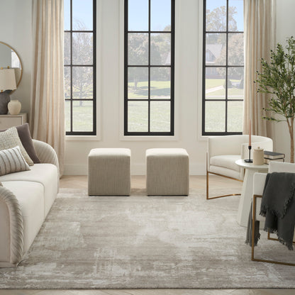 Neutral living room with cream sofa, two beige ottomans, textured rug, windows, ivory curtains, round side table & plant.