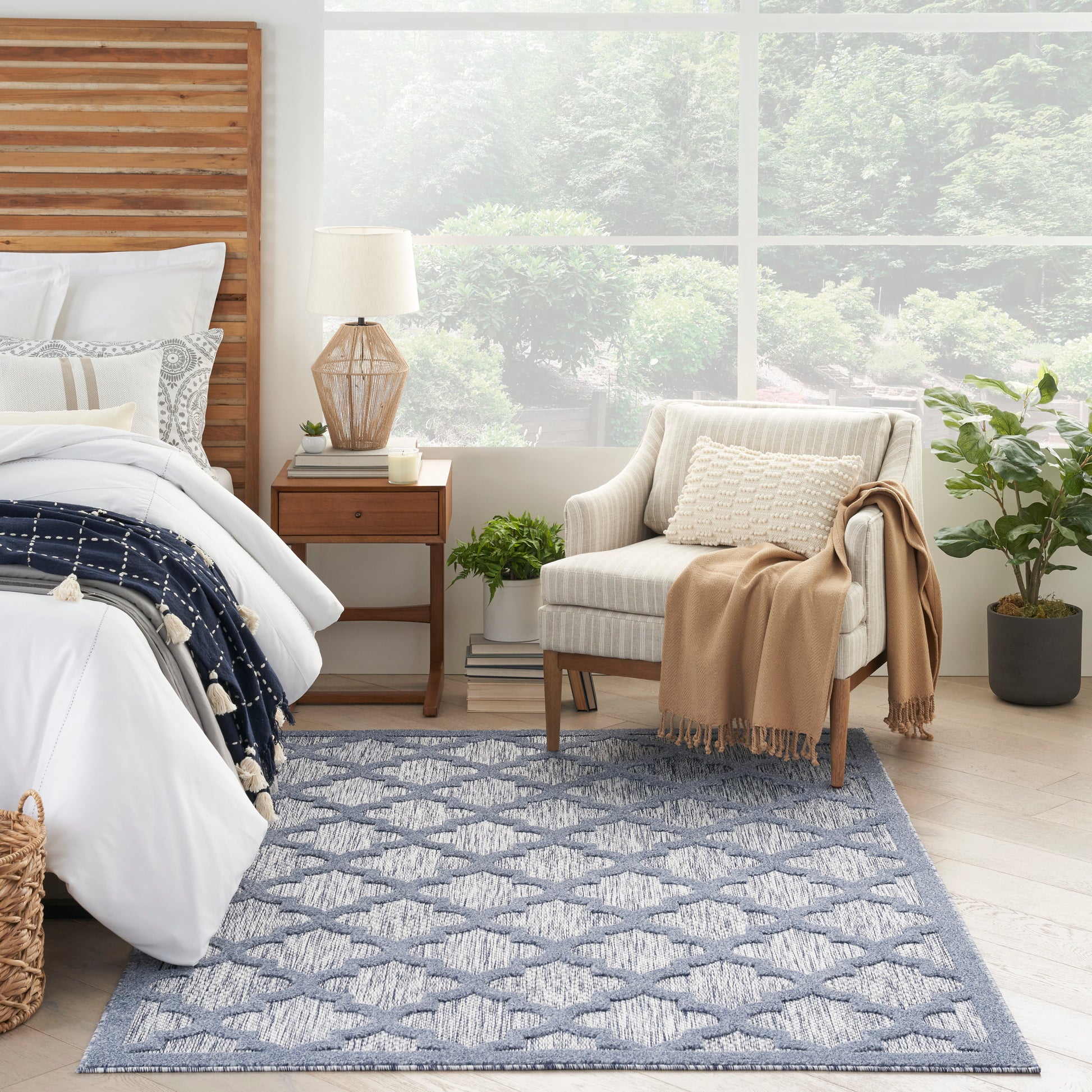 Cozy bedroom with white bedding, wooden headboard, navy throw, blue patterned rug, beige armchair, and plants.