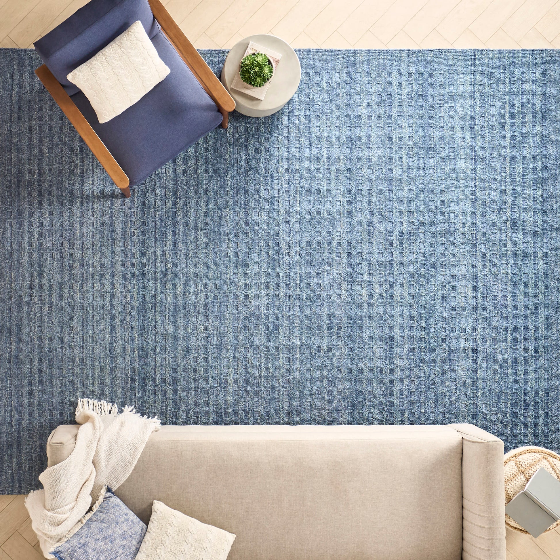 Blue textured area rug under beige sofa and navy armchair, with knit throw and round side table with plant.