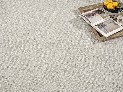 Beige woven area rug with subtle linear texture; top-right wooden tray holds open book and a bowl of lemons and grapes.