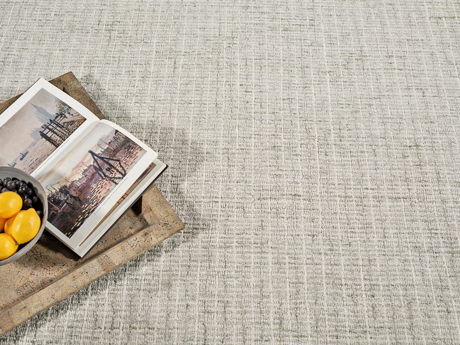 Beige textured rug with a rustic wooden tray holding an open travel book and a bowl of lemons and grapes.