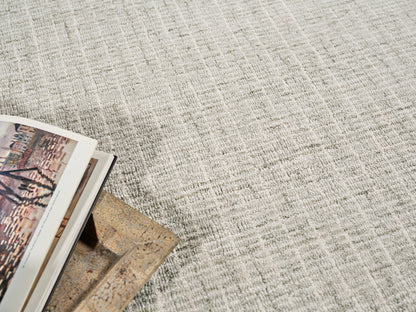 Beige textured woven rug; bottom-left corner shows magazines with colorful photos and a brown textured decorative piece.