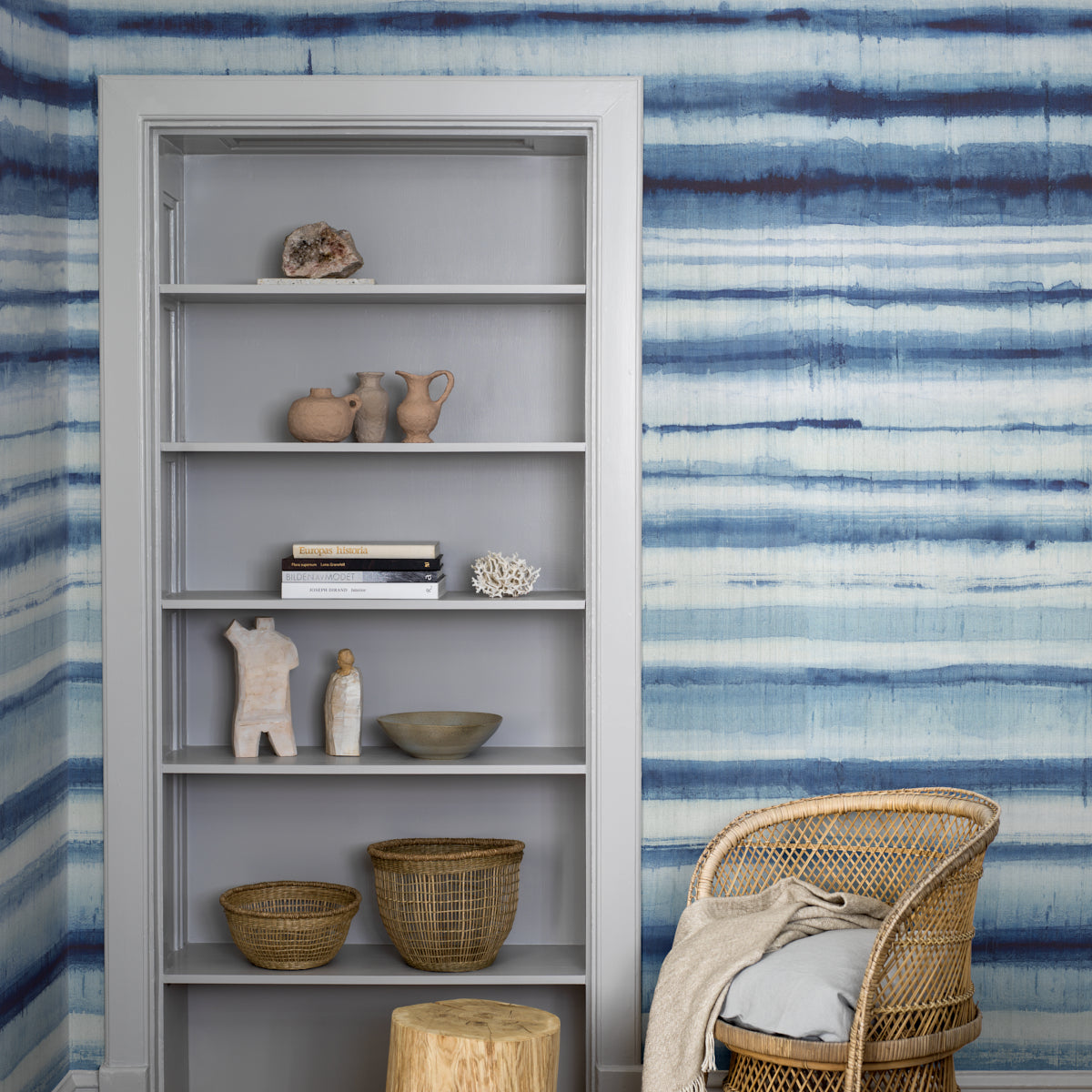 White tall bookshelf with ceramic pots, books and woven baskets against a blue striped wall; wicker chair with beige throw beside.