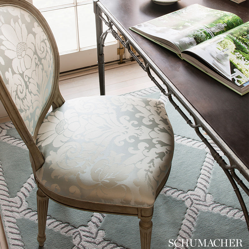Beige wooden framed chair with light blue damask upholstery beside a dark wood table & an open magazine & patterned rug.