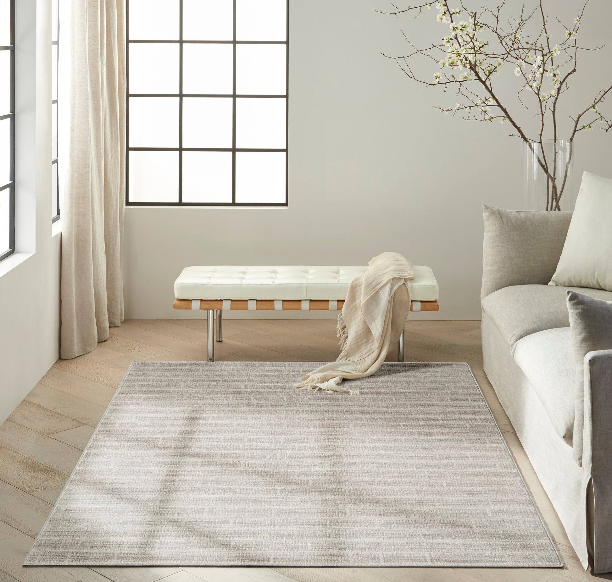 Neutral living room with a light gray-beige area rug, cream tufted bench, pale sofa, sheer curtains and a branch vase.