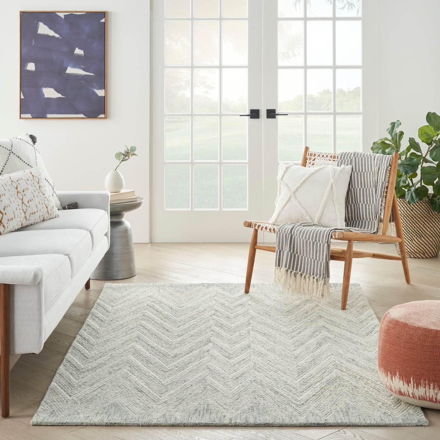 Modern living room featuring a light sofa, wooden accent chair, chevron rug, abstract blue wall art, and plants.