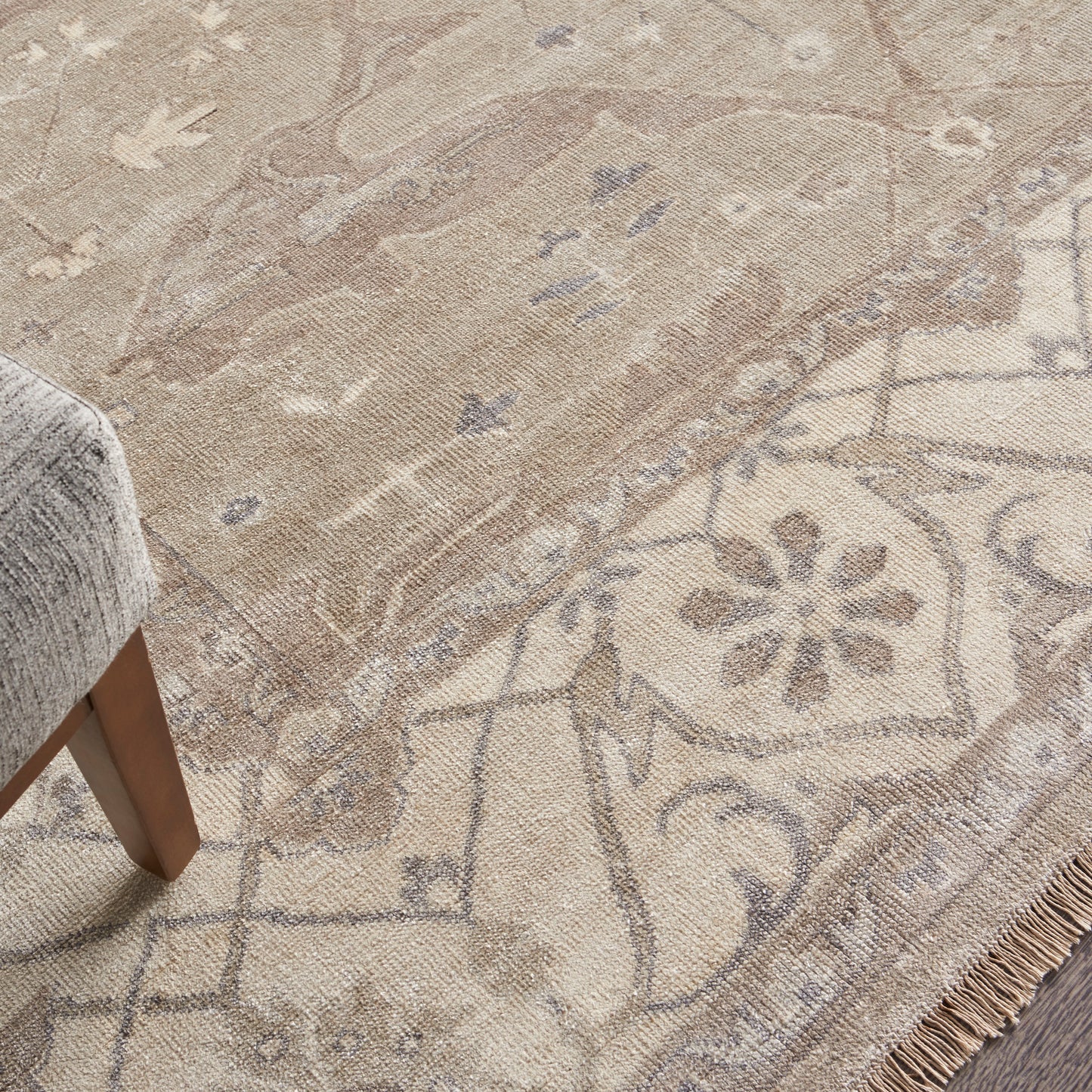 Beige oriental area rug with floral medallion motif and fringe along edge; gray upholstered furniture leg nearby.