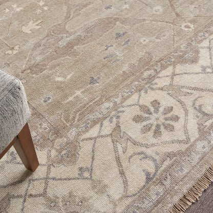 Beige oriental area rug with floral medallion motif and fringe along edge; gray upholstered furniture leg nearby.