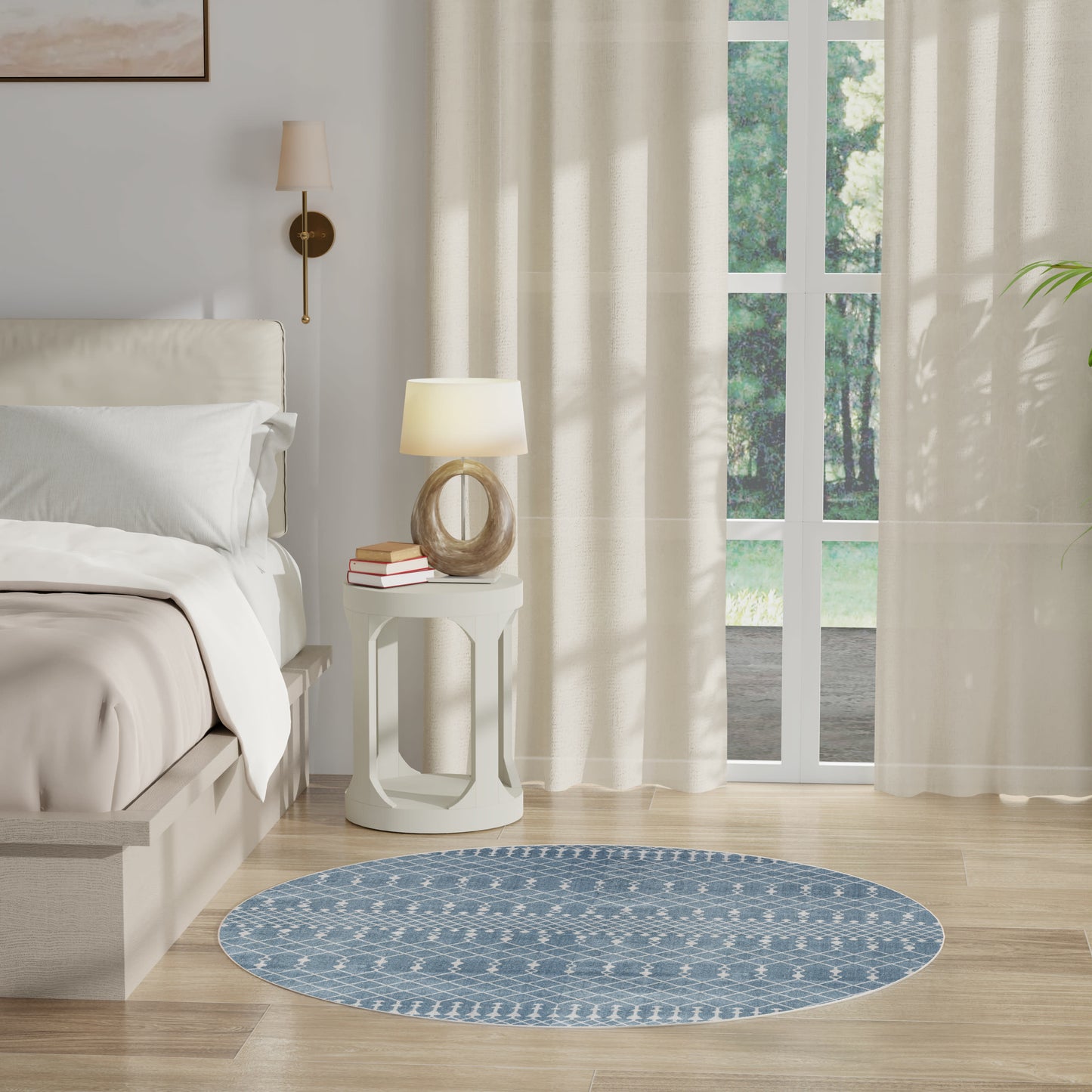 Round blue patterned rug on light wood floor beside white bed and nightstand with lamp; sheer curtains and garden view.
