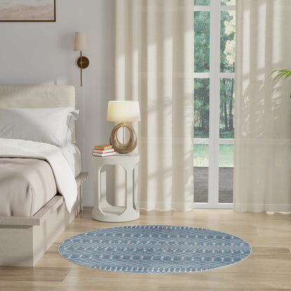 Round blue patterned rug on light wood floor beside white bed and nightstand with lamp; sheer curtains and garden view.