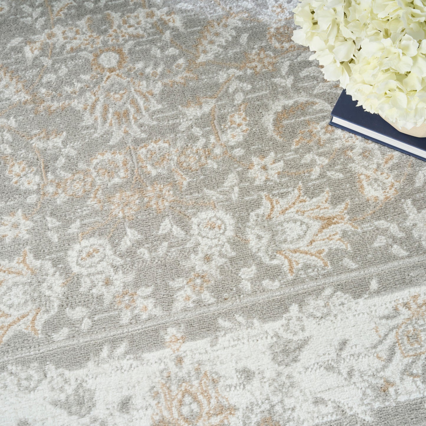 Gray floral-pattern area rug with ivory and beige motifs; white hydrangea bouquet and navy notebook in corner.