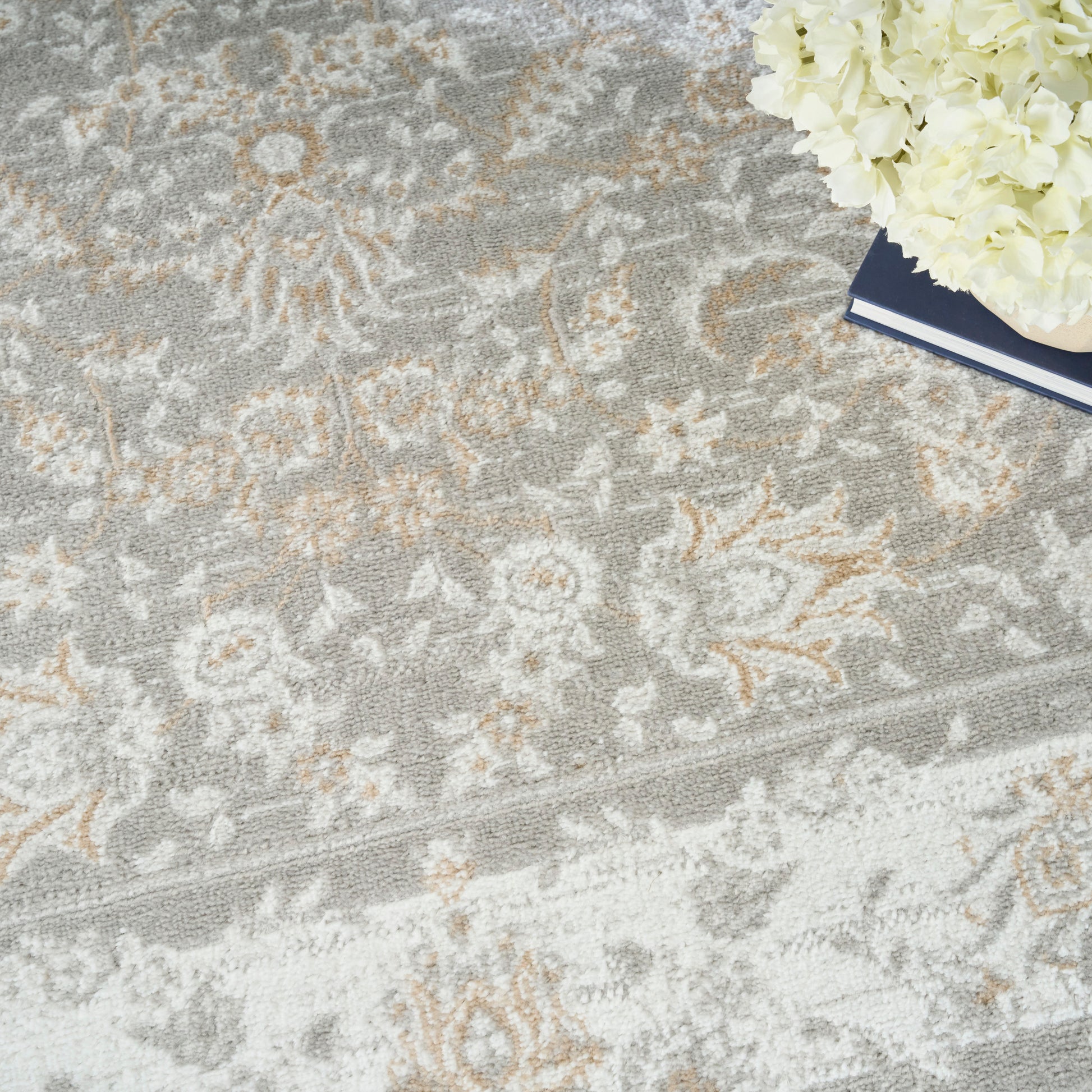 Gray floral-pattern area rug with ivory and beige motifs; white hydrangea bouquet and navy notebook in corner.