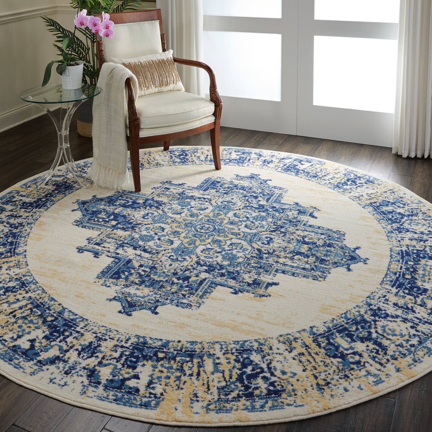 Round cream & blue distressed medallion rug in living room, paired with wooden armchair, glass side table & plant.