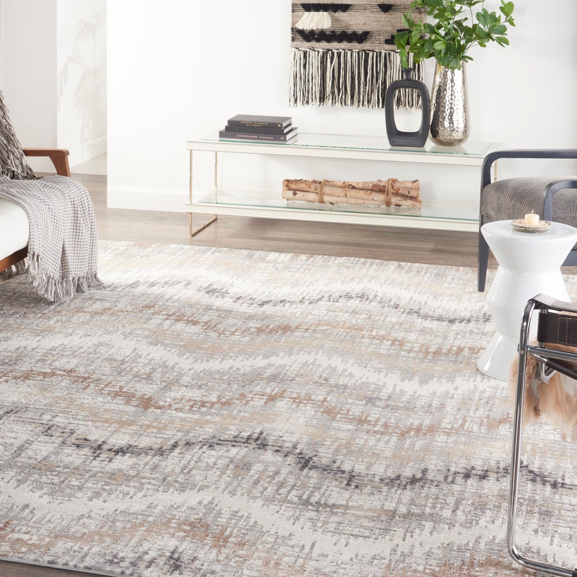 Modern living room with abstract gray-beige rug, glass console table with metal legs, black vase, plant, and sleek seating.