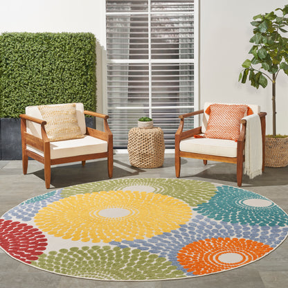 Round multicolor rug on outdoor patio with wooden-frame armchairs, beige cushions, bright pillows, wicker side table, and plants.