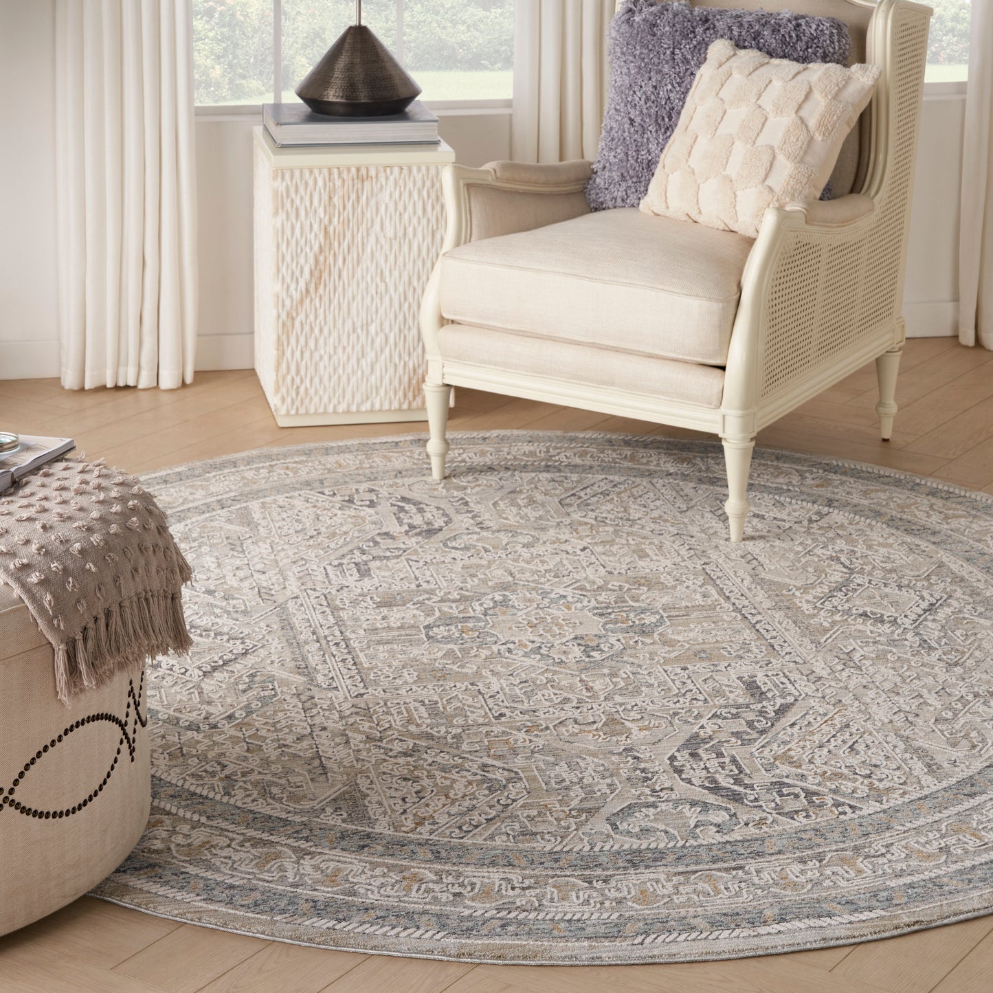 8' round gray-blue patterned rug with ivory accents, cream wicker armchair & cushions, light wood floor, lattice side table.