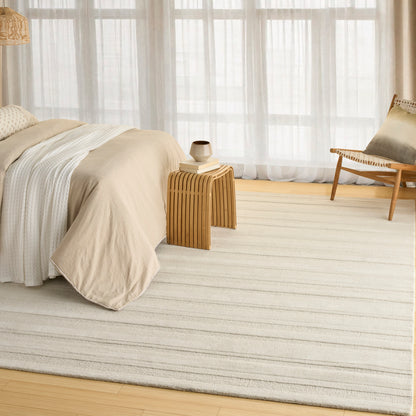 Beige linen bedding on a light striped rug with a wooden slatted nightstand and woven chair in a sunlit room.