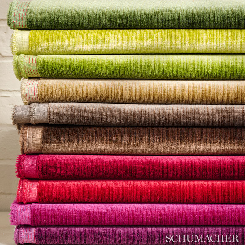 Stack of folded woven fabrics in greens, yellows, browns and pinks; textured weave, frayed edges, against a light brick wall.