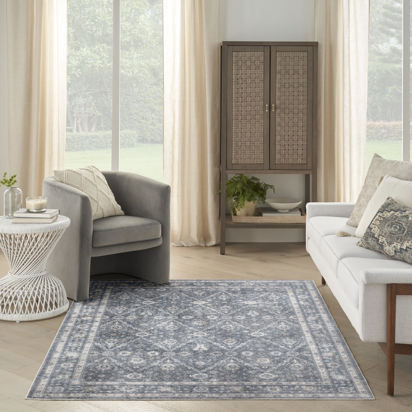 Cozy living room with light wood floors, blue patterned rug, gray armchair, white sofa, wicker cabinet, sheer curtains.