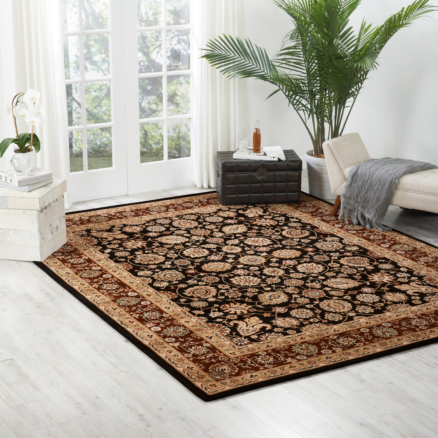 Traditional oriental area rug with black center and intricate red-gold border in a bright living room with beige sofa and plant.