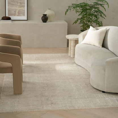 Beige curved sofa with cream cushion, round wood side table, tan chairs, light rug, potted plant in a neutral living room.