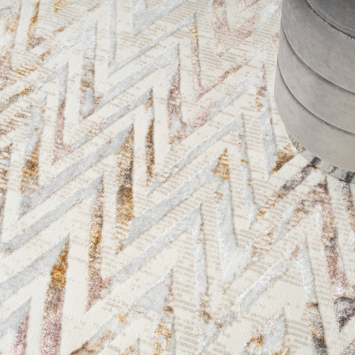 Light chevron rug with cream, beige, and grey zigzag stripes; grey round pouf shown in top-right corner.