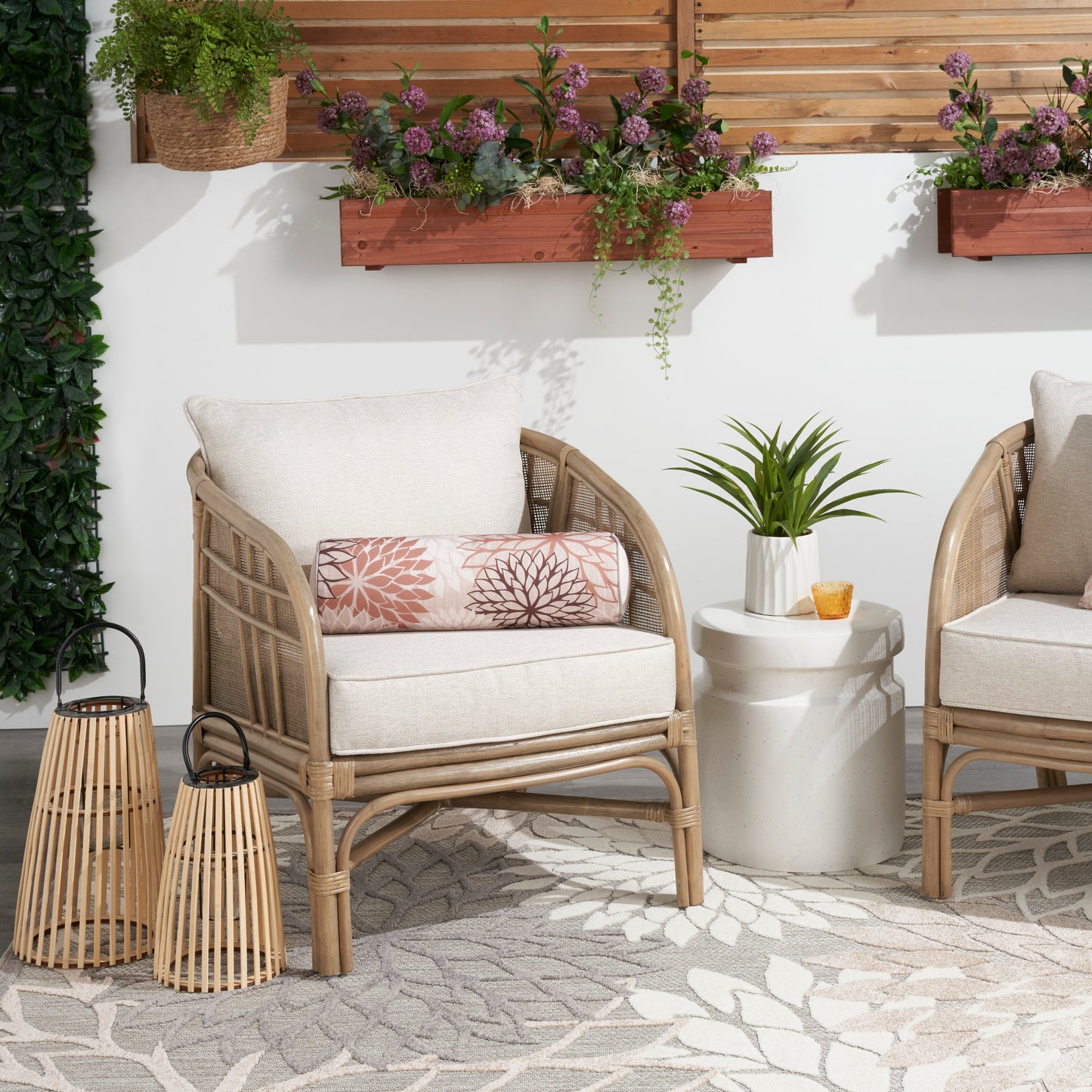 Natural tropical patterned rug on a patio with rattan chairs and beige cushions, pink lumbar pillow, white side table, lanterns and planters.