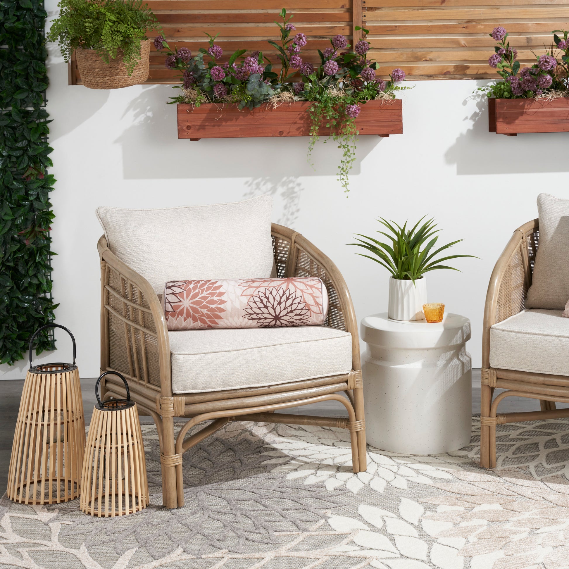 Natural tropical patterned rug on a patio with rattan chairs and beige cushions, pink lumbar pillow, white side table, lanterns and planters.