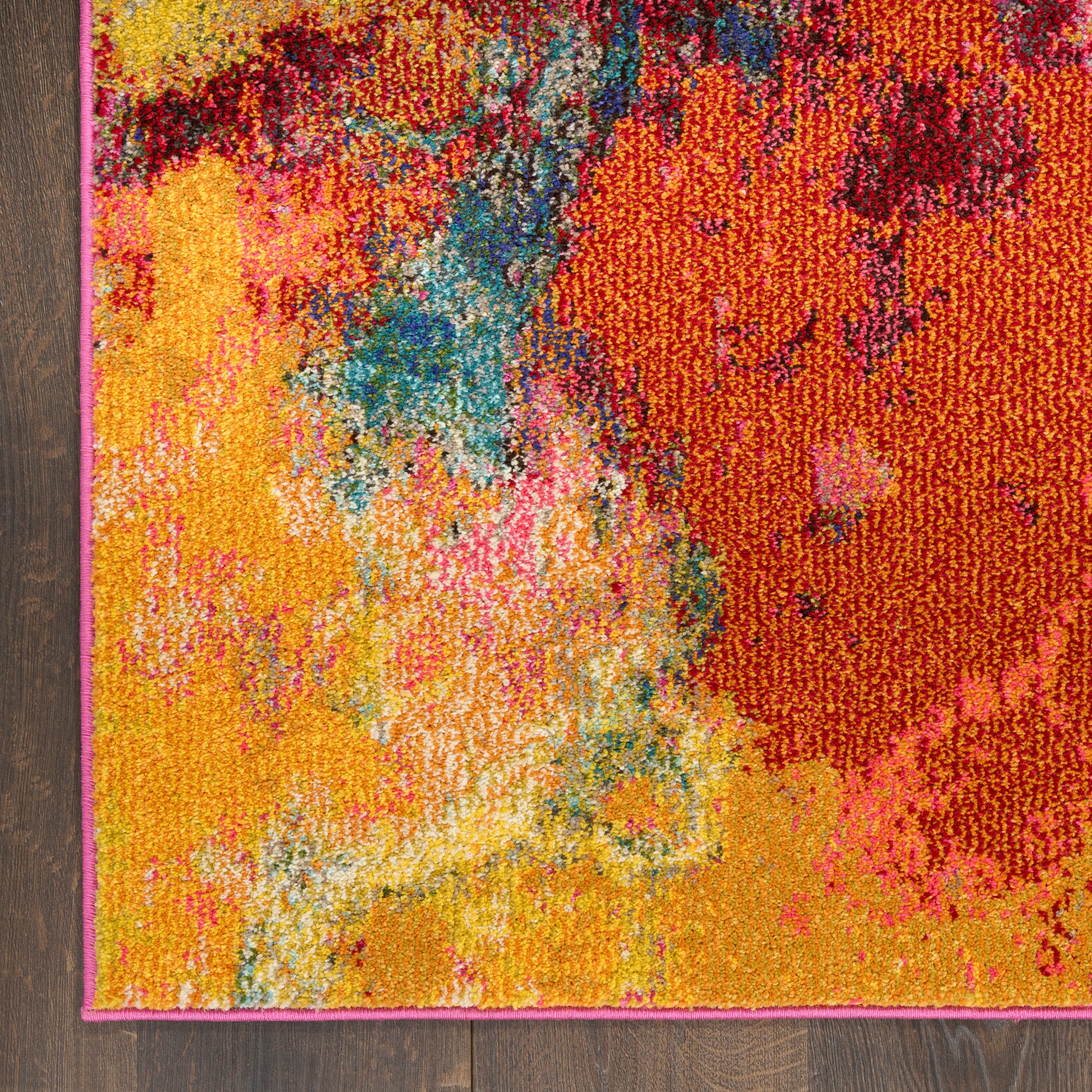 Abstract multicolor area rug with yellows, oranges, pinks and blues; pink edge binding, plush tufted texture on wood floor.