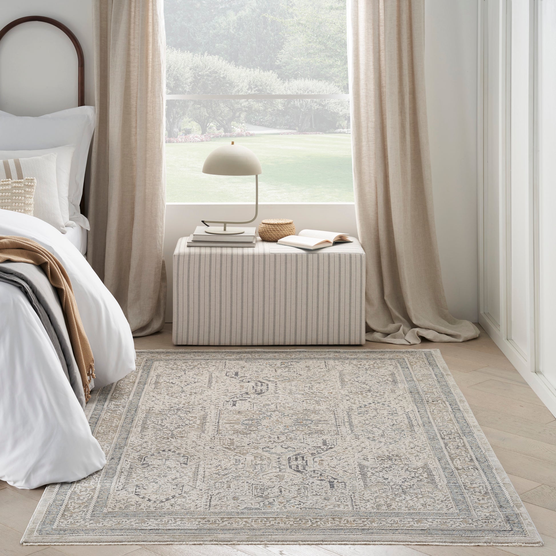 Light gray-cream patterned area rug with a medallion border in a sunlit neutral bedroom beside a striped rectangular ottoman.
