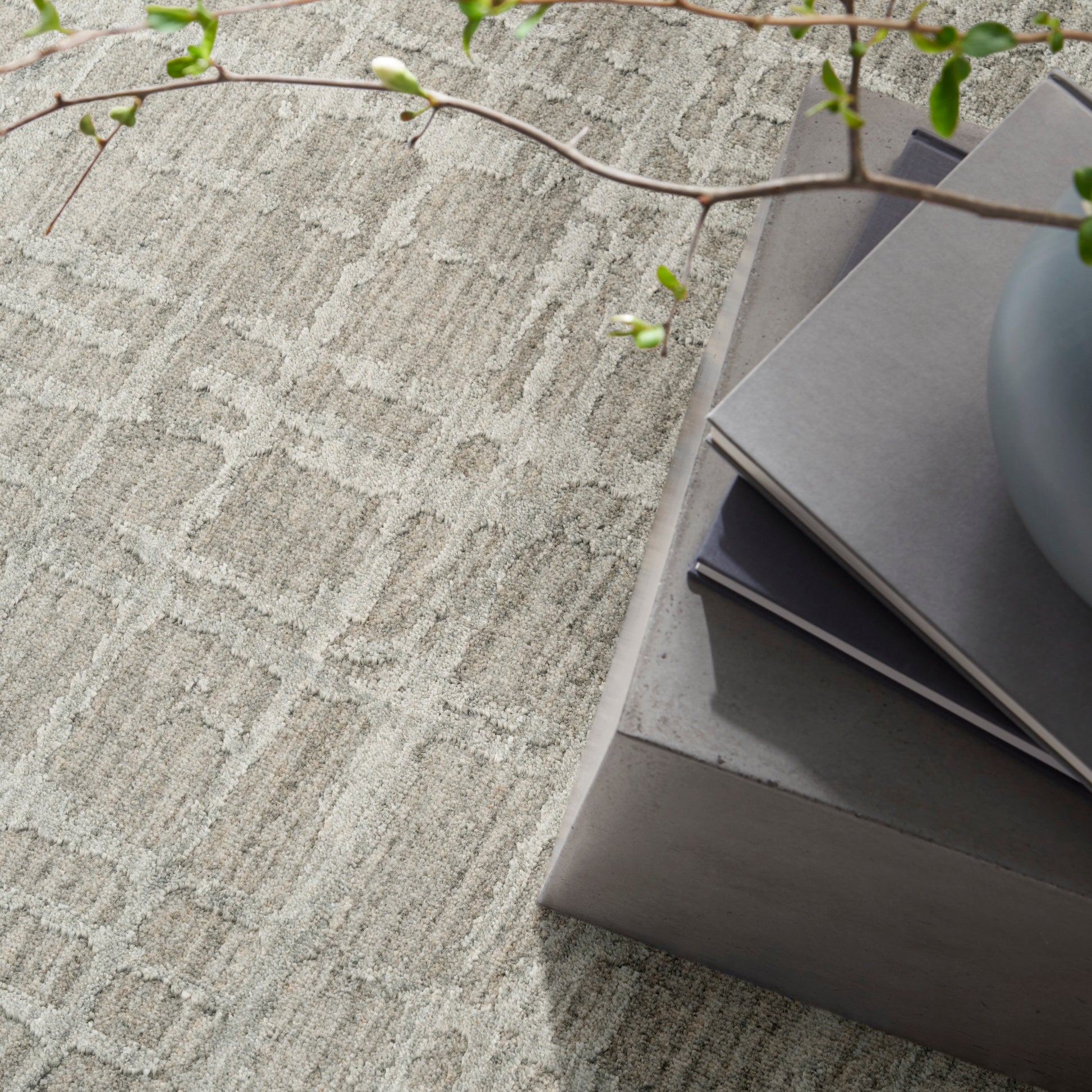 Gray textured rug with geometric pattern, stacked gray paint swatches on a concrete pedestal, green leafy branch overhead.