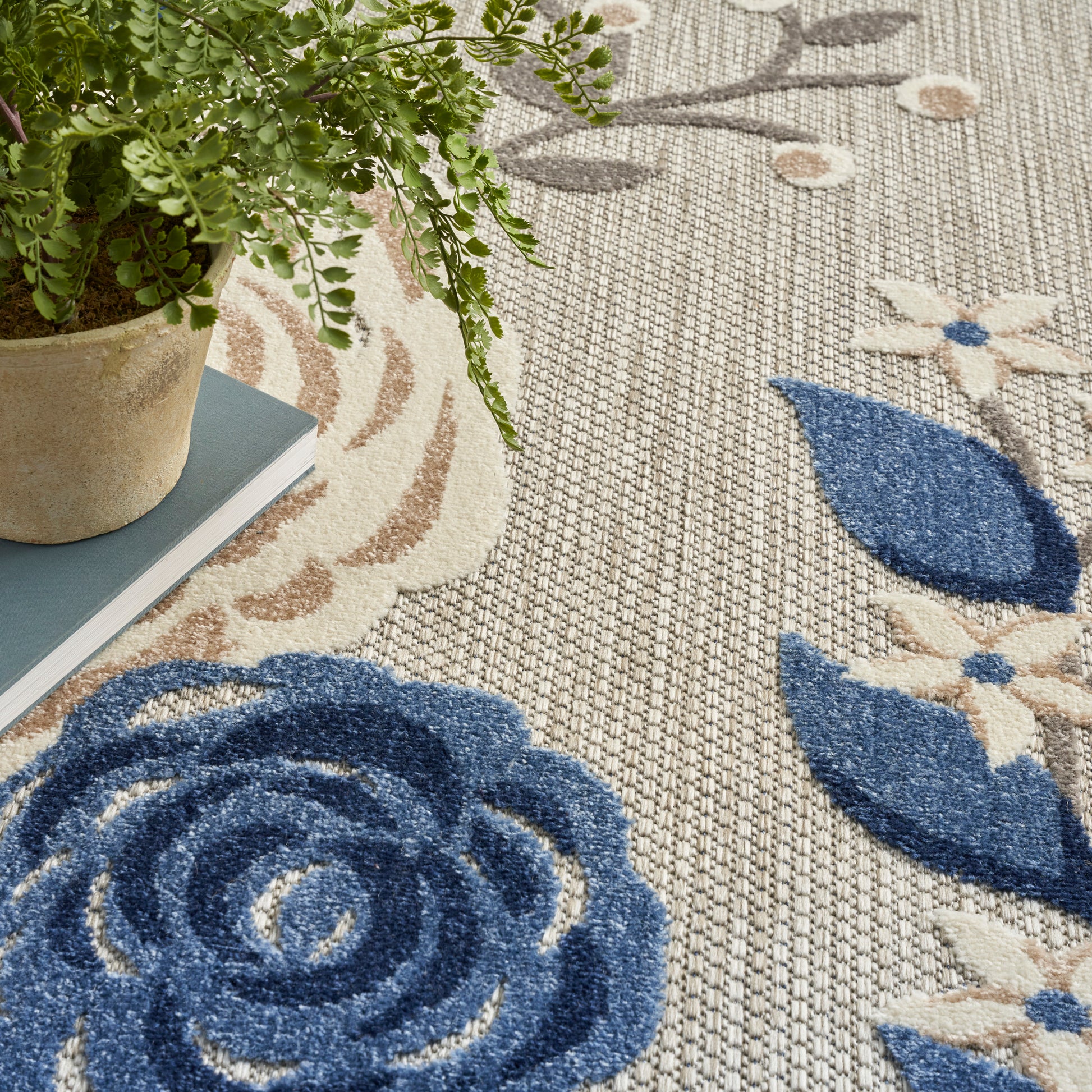 Woven beige area rug with large blue rose and leaf pattern; potted fern sits on a blue book beside it.