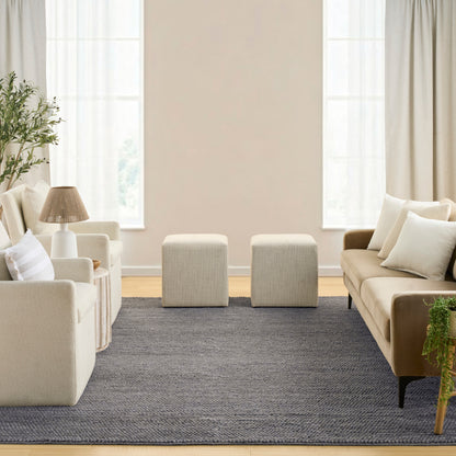 Neutral living room with cream sofas, beige armchairs, two ottomans, dark gray rug, wood floor, plants, and large windows.