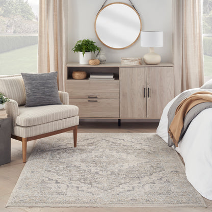 Neutral bedroom with light wood dresser, round mirror, plant, lamp, beige curtains, upholstered chair with gray pillow, and patterned rug.