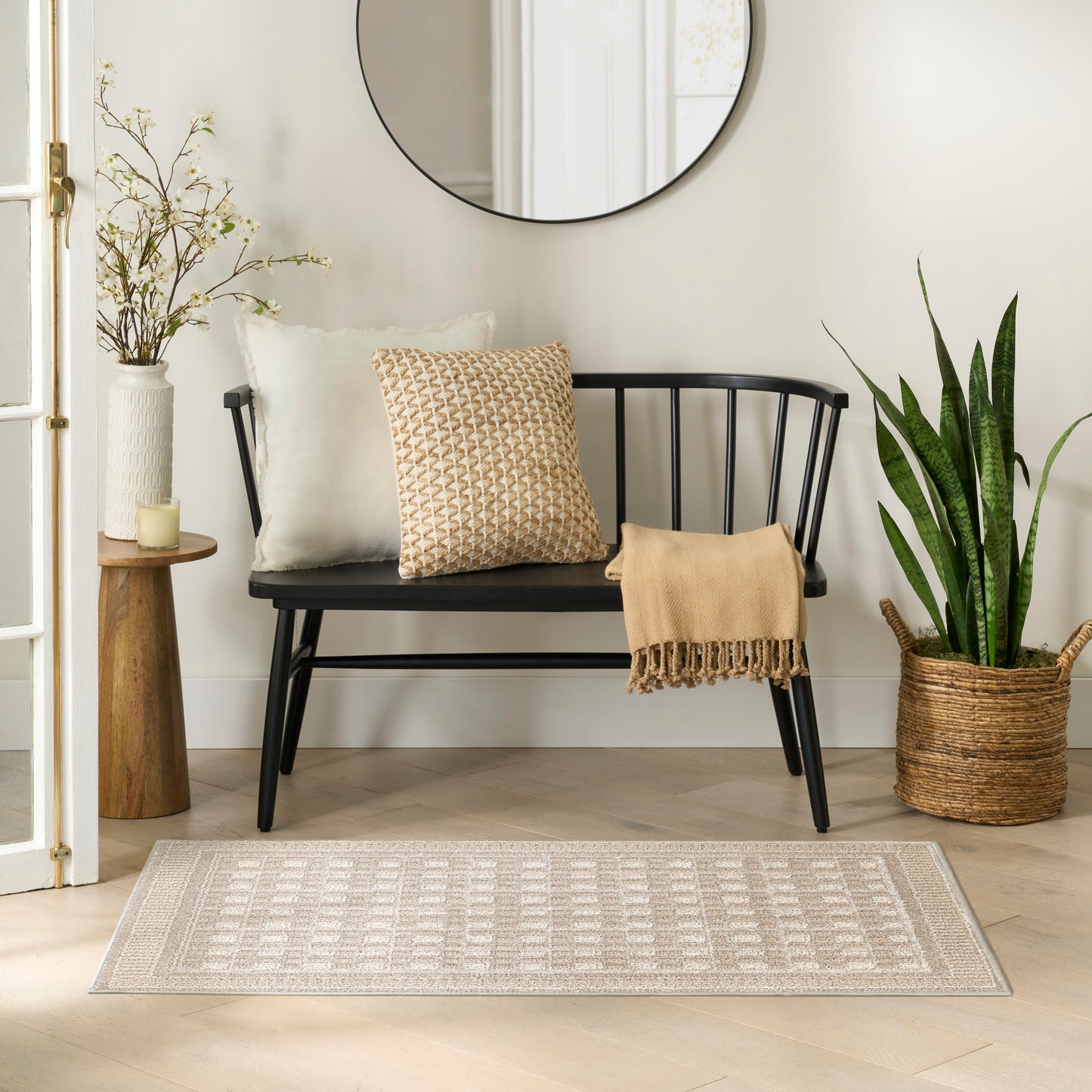 Beige modern rug with subtle pattern; part of a living room setup featuring a black wooden bench with a beige patterned cushion, tan fringed throw, round mirror and a snake plant in a woven basket.