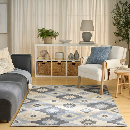 Cozy living room with light curtains, white shelves, baskets, gray sofa, cream armchair, blue pillow, and a blue-gray rug.