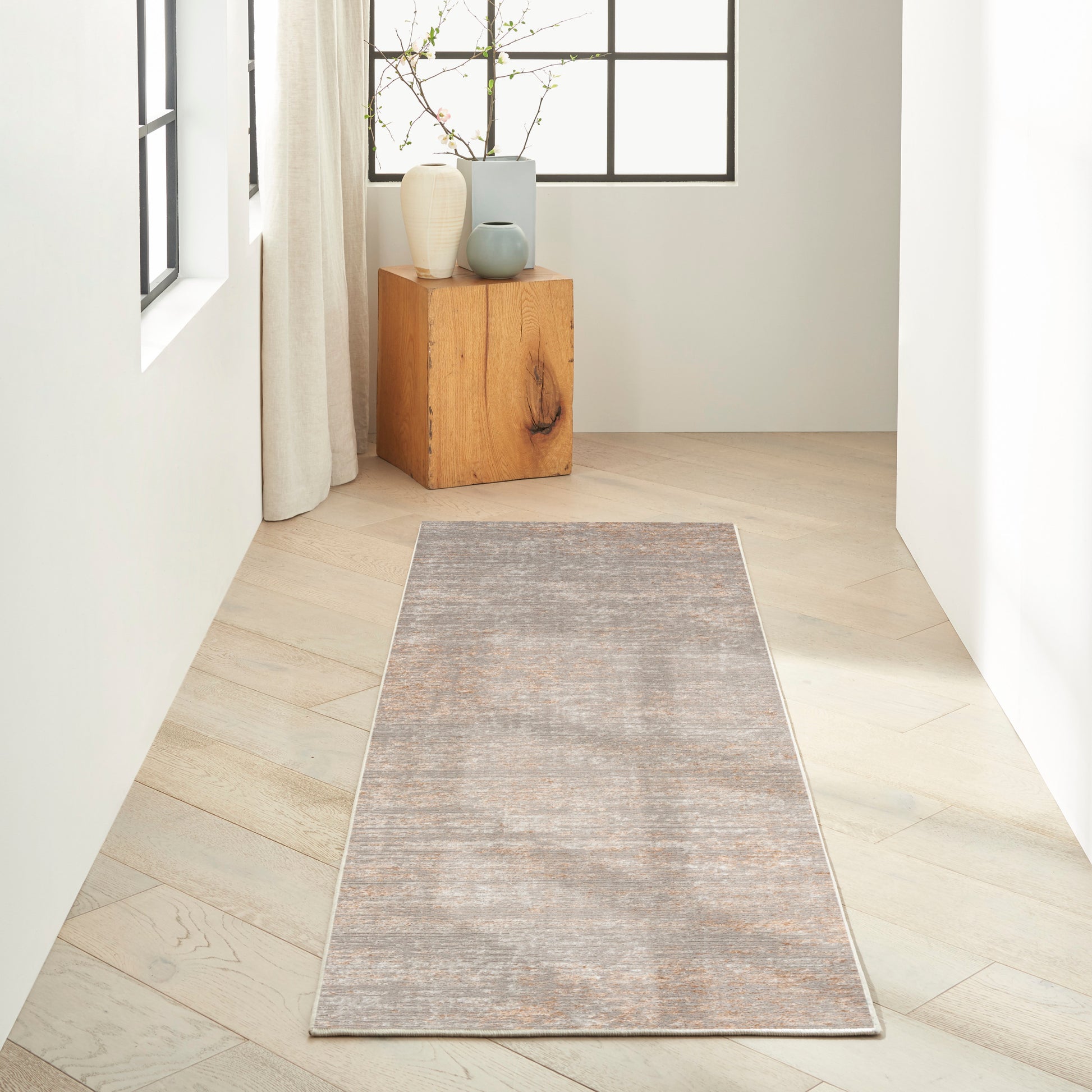 Gray-beige woven runner rug on light wood floor, with a wooden pedestal and pale vases by a window.