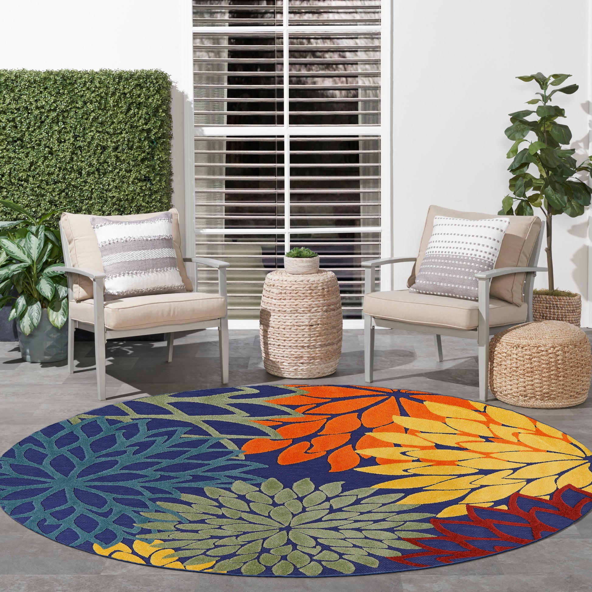 Round outdoor rug with bright blue background and multicolor floral pattern; beige cushioned patio chairs and woven stools.
