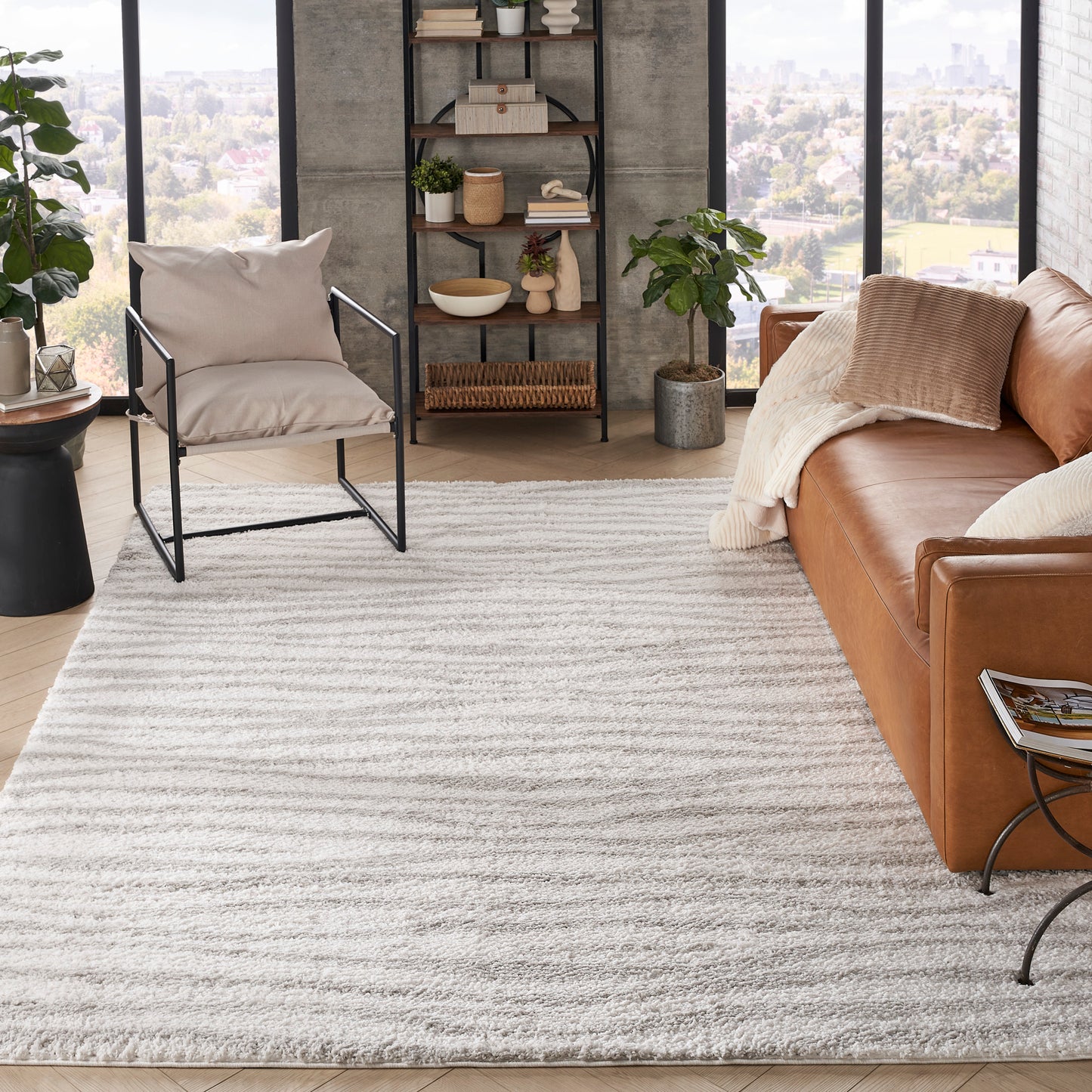 Cozy modern living room with tan sofa, beige cushions, black metal chair, plush light-gray rug, open shelf, plants, view.
