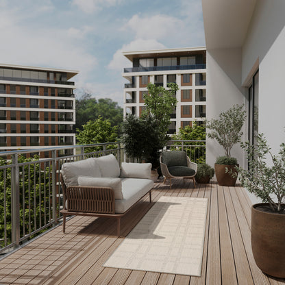 Sunlit balcony with cream-cushioned sofa, dark wicker chair, beige rug, potted plants, wooden deck, and buildings beyond.