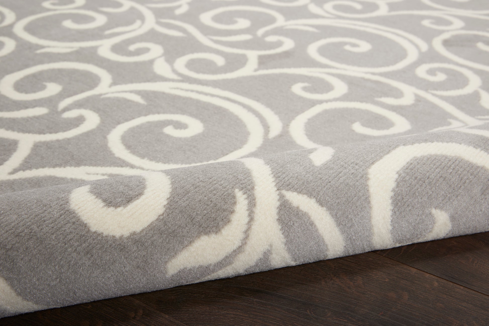 Close-up of gray rug with white swirling scroll pattern on dark wood floor.