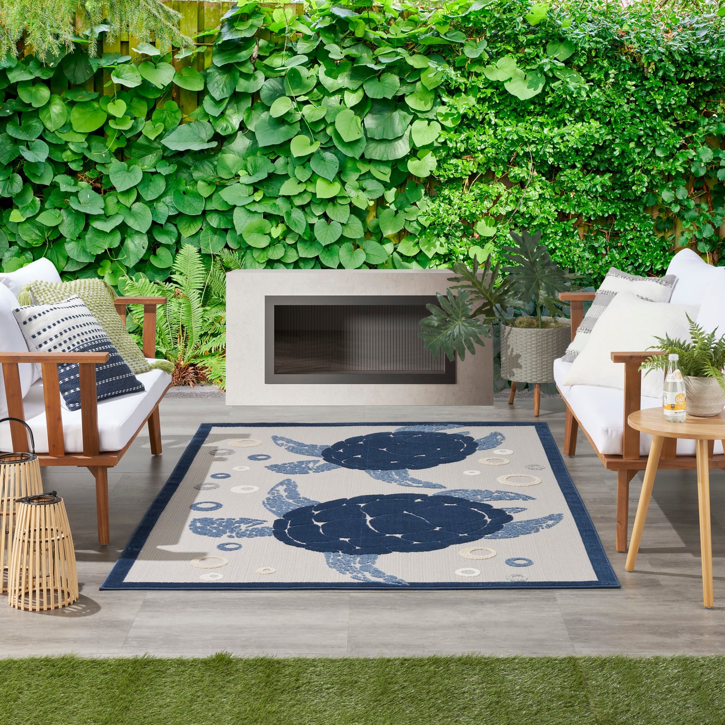 Outdoor patio seating around a beige rug with navy circular shapes, modern rectangular fireplace, and lush green hedges.