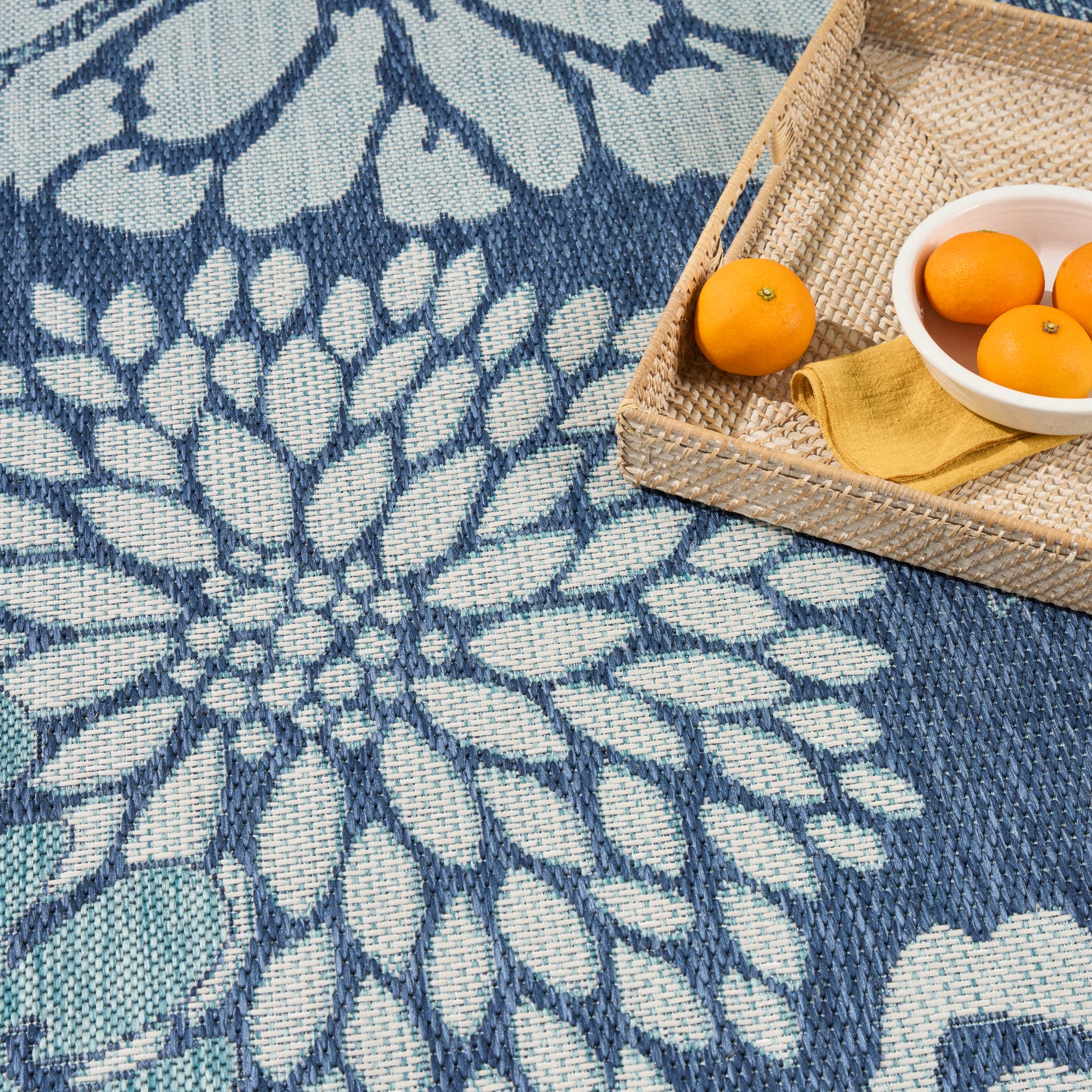 Blue and teal woven rug with a large floral pattern; wicker tray holding oranges, a white bowl, and a mustard napkin.