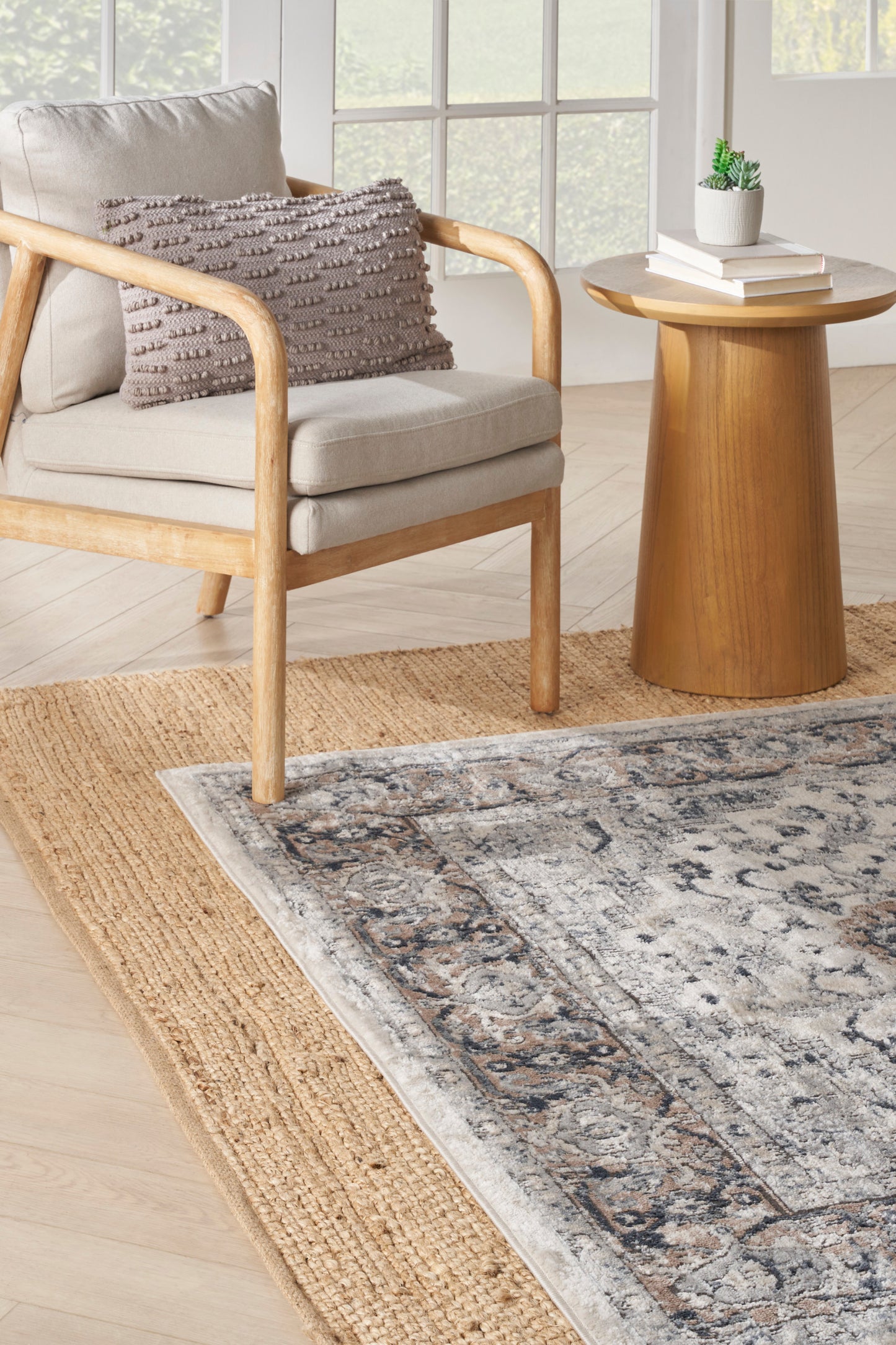 Light wood armchair with beige cushions and textured taupe pillow, round side table, plant, on jute and blue-gray rug.