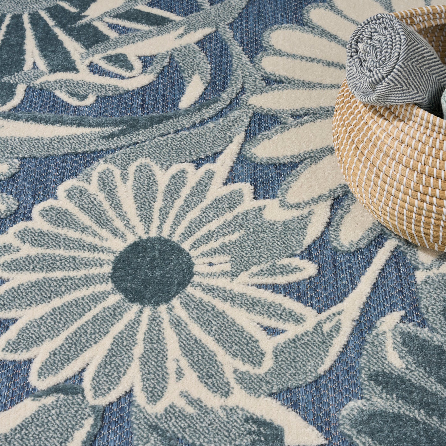 Blue rug with large white daisy floral motif; woven texture & a round wicker basket with a rolled striped towel.