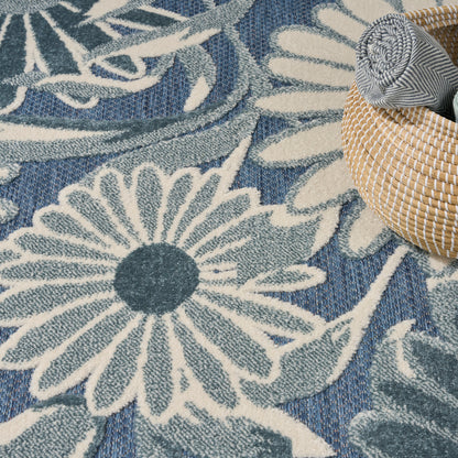 Blue rug with large white daisy floral motif; woven texture & a round wicker basket with a rolled striped towel.
