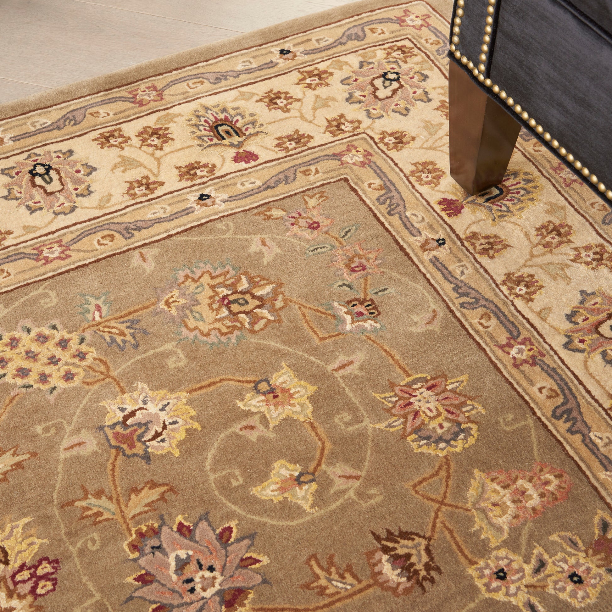 Beige oriental area rug with pastel floral motifs and blue-bordered edges on a wood floor beside a nailhead-studded chair leg.