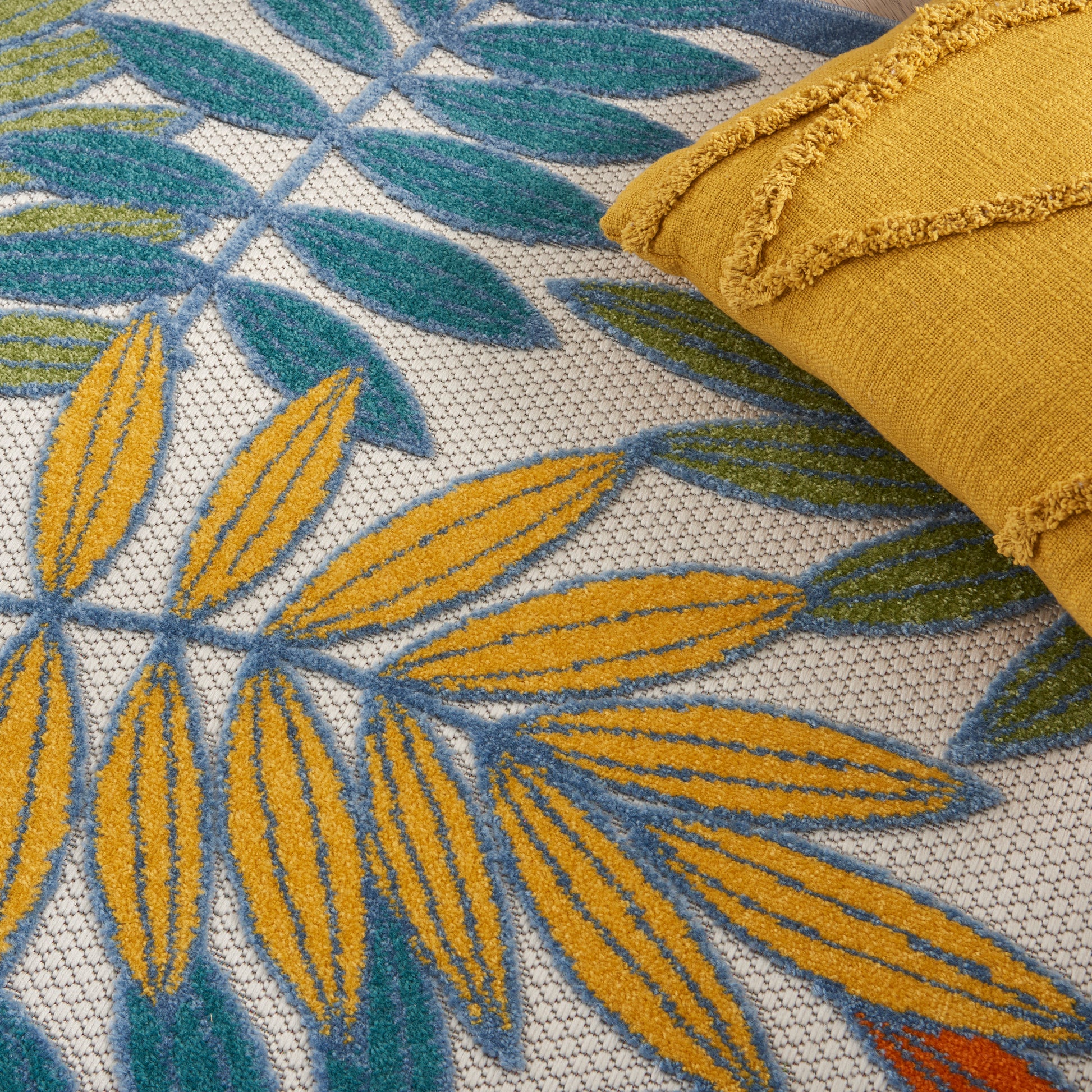 Yellow and teal tropical leaf-pattern fabric with a textured weave; mustard fringe-edged roll resting on it.